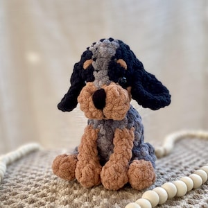May include: A handmade crochet dog plushie in a sitting position. The dog features a black and gray head and ears, a tan muzzle and paws, and a gray and tan body. The plushie is sitting on a textured surface.