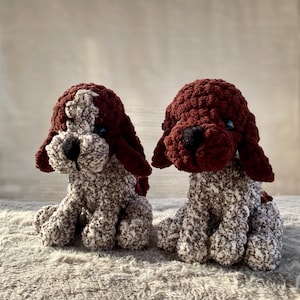 May include: Two handmade crochet dog stuffed animals. The dogs have brown and white speckled bodies, dark brown heads and ears, and black noses. The dogs are sitting on a gray surface.