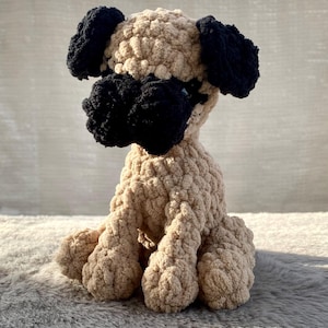 May include: A handmade, plush pug dog in a seated position. The dog features a tan body, black ears and snout, and is made of a soft, textured yarn. The toy is designed to resemble a pug breed.