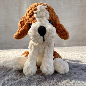 May include: A handmade crochet dog plushie in a sitting position. The dog features brown and white yarn, with black eyes and nose. The plush toy is soft and fluffy, perfect for a child's room or as a decorative item.