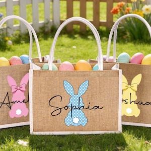 Könnte beinhalten: Drei Ostereinkaufstaschen aus Jute mit weißen Griffen und Seiten, jede mit einem Hasenmuster und einem Namen. Die Taschen sind mit bunten Ostereiern gefüllt. Der Hintergrund umfasst einen weißen Zaun und Frühlingsblumen.