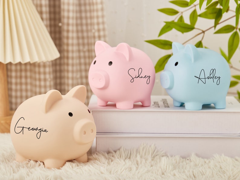 Pu&ograve; includere: Tre salvadanai a forma di maiale, uno rosa, uno blu e uno beige, con nomi personalizzati: Georgia, Sidney e Ashley. I salvadanai sono su una superficie bianca.