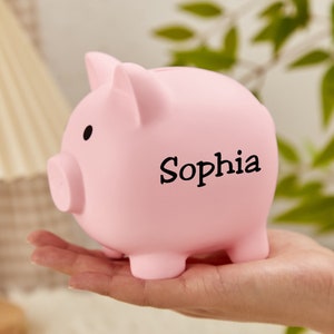 Pu&ograve; includere: Salvadanaio a forma di maiale rosa con un'iscrizione nera che dice "Sophia".