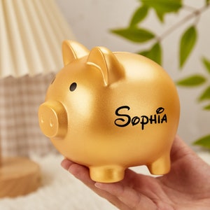 Pu&ograve; includere: Salvadanaio a forma di maiale dorato con il nome "Sophia" scritto in lettere nere.