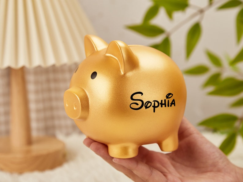 Pu&ograve; includere: Salvadanaio a forma di maiale dorato con il nome "Sophia" scritto in lettere nere.