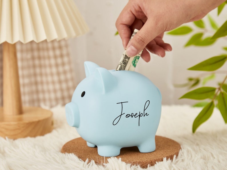 Pu&ograve; includere: Un salvadanaio di colore azzurro chiaro con il nome "Joseph" scritto su di esso. Una mano sta mettendo una banconota da un dollaro nella fessura.