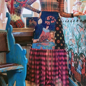 Könnte beinhalten: Eine Frau trägt einen langen Rock mit Patchwork-Design. Der Rock hat ein blaues Feld mit einer sitzenden Figur, ein schwarzes Feld mit orangefarbenen Kreisen und einen rosa-schwarz karierten unteren Abschnitt. Sie trägt auch ein langärmliges Oberteil mit Karomuster.