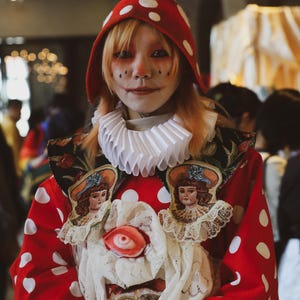 May include: A person wearing a red and white polka dot costume with a white ruffle collar and a red mushroom hat. The costume features appliques of dolls and lace.