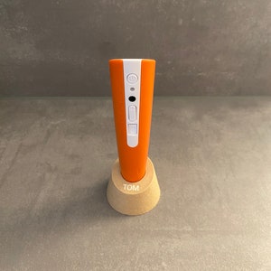 Könnte beinhalten: Ein orangefarbenes und weißes elektronisches Gerät mit Steuertasten, das in einem hellen Holzständer steht. Auf dem Ständer steht der Name "TOM". Das Gerät ist zylindrisch und scheint ein Handwerkzeug zu sein.