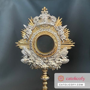 May include: A gold and silver monstrance with a round, clear glass center. The monstrance has a crown on top and two angels on either side. The base is decorated with intricate details.