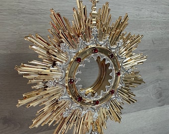 May include: A gold monstrance with a silver base and a red jeweled halo. The monstrance is decorated with a cross at the top and a sunburst design.