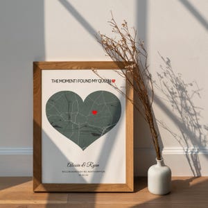May include: A framed print featuring a heart-shaped map in shades of gray, with a red heart marking a specific location. The text "THE MOMENT I FOUND MY QUEEN" is at the top, and names and a date are at the bottom. The frame is made of wood.