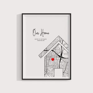 May include: Black framed art print. The print features a house-shaped map with a red heart marking a location. The words "Our Home" are written above the map, with coordinates and a date below. The print is in black and white.