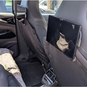 May include: Interior view of a car with a tablet mounted on the back of a front seat. The tablet is in a black frame and is attached to the seat with a metal bracket. The car's interior is black and gray, with a child car seat visible.