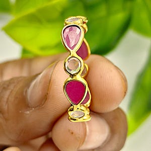 Könnte beinhalten: Ein goldener Ring mit abwechselnd tropfenförmigen Rubinen und klaren Edelsteinen. Das Design des Rings ist ein durchgehendes Band aus Edelsteinen, das einen funkelnden und eleganten Look erzeugt. Der Ring wird vor einem verschwommenen grünen Hintergrund gehalten.