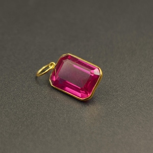 May include: A rectangular pendant featuring a faceted, deep pink gemstone set in a gold-toned metal frame. The pendant includes a small, circular gold-toned bail attached to one side, designed for suspension on a chain or cord.