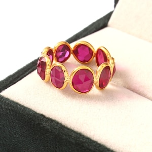 Puede incluir: Un anillo dorado con una serie de piedras preciosas de color rojo rubí, de corte ovalado. Las piedras están engastadas en biseles individuales, creando una banda continua. El anillo se muestra en una caja de joyería verde oscuro.