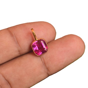 Puede incluir: Un colgante de tono dorado con una piedra preciosa de color rosa vibrante y corte cuadrado. El colgante tiene una sencilla anilla de metal en la parte superior para su sujeción. La piedra tiene una superficie facetada que refleja la luz, mostrando un tono magenta intenso y un acabado pulido.
