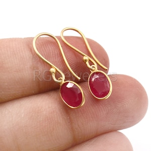 May include: A pair of gold earrings with red oval-shaped gemstones. The earrings have a simple, elegant design.