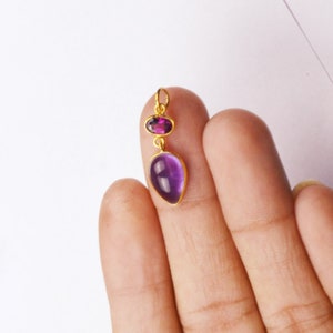 May include: A gold pendant with a purple amethyst gemstone and a smaller purple garnet gemstone.