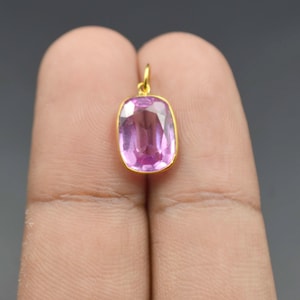 May include: A cushion-cut pink gemstone pendant set in a thin gold bezel frame. The gemstone features a faceted surface that reflects light, and a small gold jump ring is attached to the top of the setting for hanging on a chain or cord.