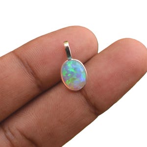 May include: An oval-shaped pendant featuring a polished, iridescent opal gemstone set in a smooth silver-toned metal bezel. The opal displays vibrant flashes of green, blue, and white light across its surface, suspended from a small silver bail.
