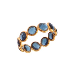 Puede incluir: Un anillo dorado con piedras preciosas azules ovaladas y facetadas. Las piedras están dispuestas en una banda continua. El diseño del anillo es sencillo y elegante. El anillo tiene un diámetro de aproximadamente 1,9 cm y es adecuado para cualquier ocasión.