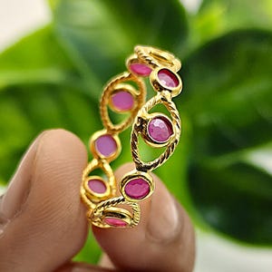 Peut inclure: Une bague dorée ornée d'une série de pierres précieuses roses ovales. La bague présente un motif torsadé entre chaque pierre précieuse. La bague est présentée sur un fond vert flou.