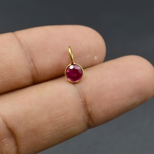 Op de afbeelding: Een kleine, ronde, gefacetteerde dieproze edelsteen gezet in een gepolijste gouden zetting. De hanger is voorzien van een delicaat gouden ringetje aan de bovenkant, ontworpen om aan een ketting of koord te hangen, met een verfijnde uitstraling.
