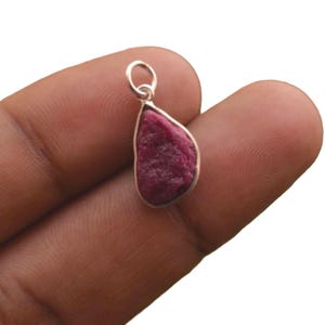 May include: A teardrop-shaped pendant featuring a raw, deep pinkish-red gemstone set in a simple silver-toned metal bezel. The pendant includes a small circular silver-toned jump ring at the top for attachment to a chain or cord.