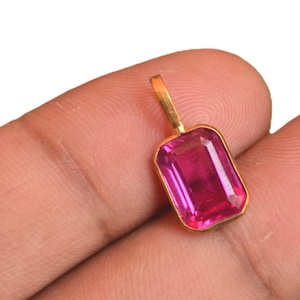 May include: A rectangular pendant featuring a vibrant, faceted pink gemstone set in a polished gold-toned metal frame. The pendant includes a small, integrated gold-toned bail at the top, designed for attachment to a chain or cord for jewelry wear.