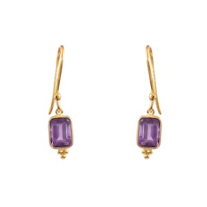 Puede incluir: Un par de pendientes de oro con piedras preciosas de amatista púrpura. Los pendientes tienen un diseño sencillo con una pequeña piedra rectangular que cuelga de una delicada cadena de oro.