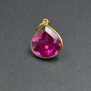 Pode incluir: Um pingente em forma de gota com uma pedra preciosa facetada de cor rosa profundo, engastada numa fina moldura de metal dourado. O pingente inclui um pequeno anel de junção circular dourado no topo para fixação, com superfície polida.