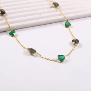 May include: A gold-colored necklace featuring a delicate chain adorned with alternating green and gray gemstones. The gemstones are cut into various shapes, including triangles and ovals. The necklace is displayed on a white surface.