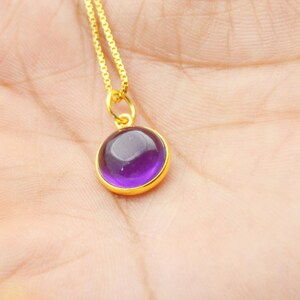 Peut inclure: Un collier doré avec un pendentif rond en pierre précieuse violet foncé. Le pendentif est serti dans une lunette dorée et suspendu à une fine chaîne dorée. La pierre précieuse est d'un violet riche et vibrant.
