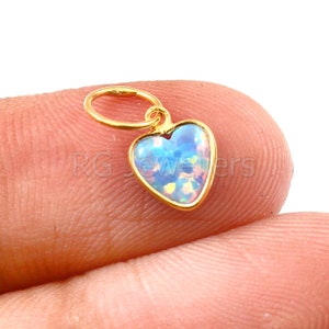 Pode incluir: Um amuleto de ouro em forma de coração com uma pedra de opala azul. O amuleto está em um dedo.