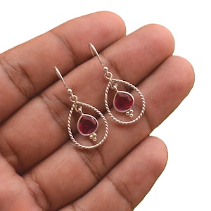 May include: A pair of silver drop earrings featuring a twisted metal teardrop frame. Each earring contains a single faceted red gemstone suspended in the center, accented with three small silver beads at the base of the stone and a hook wire.