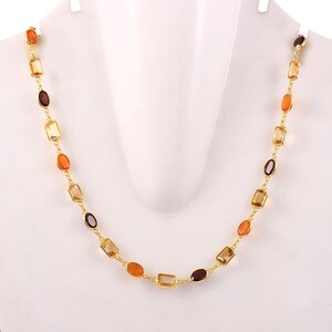 Könnte beinhalten: Eine goldfarbene Halskette mit einer Reihe farbiger Edelsteine in Orange-, Gelb- und Brauntönen. Die Edelsteine sind in einem linearen Muster angeordnet, wodurch ein zartes und elegantes Design entsteht. Die Halskette wird auf einer weißen Schaufensterpuppe präsentiert.