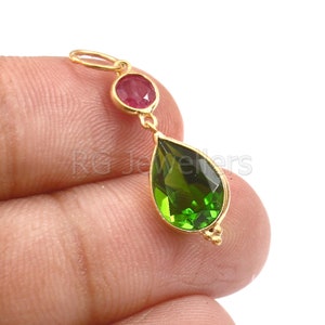 May include: A gold pendant featuring a round, red gemstone and a large, pear-shaped, green gemstone. The pendant has a small loop at the top for a chain and decorative accents at the bottom of the green gemstone.