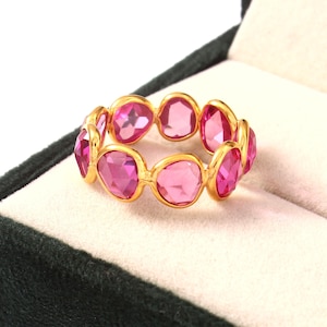 May include: A gold ring with a series of pink gemstones. The oval-shaped gemstones are set in a gold band. The ring is displayed on a white surface inside a dark green jewellery box.
