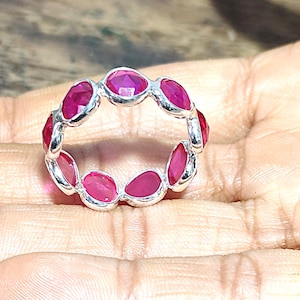 May include: A silver ring featuring a continuous band of oval-cut, vibrant red gemstones. The gemstones are set closely together, creating a full circle of color. The ring is displayed on a neutral background.