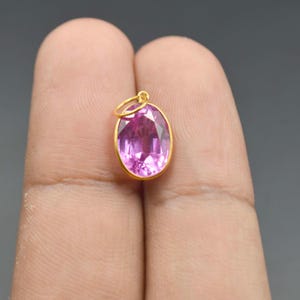 Puede incluir: Un colgante de piedra preciosa rosa de forma ovalada y tallada, engastado en un fino bisel de metal de color dorado. El colgante cuenta con una pequeña anilla circular de color dorado en la parte superior para colgarlo de una cadena.