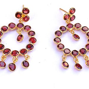 Könnte beinhalten: Ein Paar goldfarbene Ohrringe mit einem floralen Design oben und einer kreisförmigen Anordnung roter Edelsteine. Zusätzliche rote Edelsteine hängen von dem kreisförmigen Design herab.