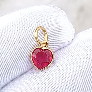 May include: A gold heart-shaped pendant with a bright red gemstone. The pendant has a small loop at the top for attaching to a chain or bracelet. The gemstone is faceted, catching the light and creating a sparkling effect.