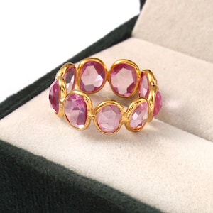 May include: A gold ring with a sequence of oval-cut pink gemstones. The gemstones are set in a gold band, forming a continuous pattern. The ring is presented in a dark green velvet jewellery box.
