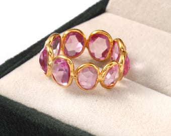 Könnte beinhalten: Ein goldener Ring mit einer Reihe von oval geschliffenen, rosa Edelsteinen. Die Edelsteine sind in einem goldenen Band gefasst, wodurch ein durchgehendes Muster entsteht. Der Ring wird in einer dunkelgrünen Samt-Schmuckschatulle präsentiert.