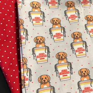 May include: A white fabric with a repeating pattern of brown dogs in a cup of noodles. The text "Cup Noodles" is printed on the cup. The background is a light gray with small red hearts.