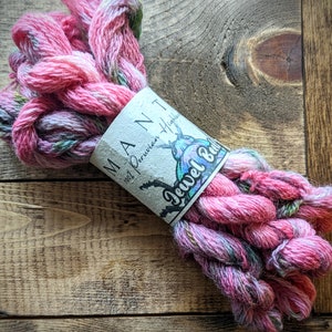 May include: A skein of pink and white yarn with green flecks. The yarn is labeled "Mant 100% Peruvian Highland Jewel Blend".