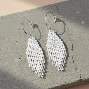 Op de afbeelding: Een paar witte kralen oorbellen met een franjedesign. De oorbellen zijn druppelvormig en hangen aan zilverkleurige hoepels. De oorbellen worden gepresenteerd op een grijze ondergrond.