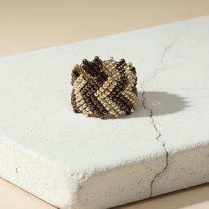 May include: A beaded ring in a zig-zag pattern, featuring alternating rows of gold and dark brown beads. The ring is set against a neutral background, showcasing its intricate design and craftsmanship. This handmade ring is a unique piece of jewelry.